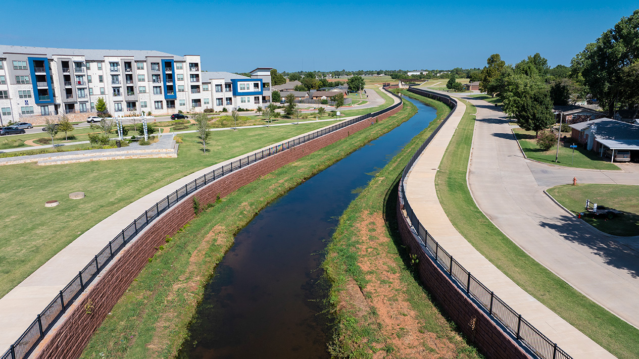 Aerial view of a landscaped drainage channel with walking paths and black metal fencing, adjacent to a multi‑story apartment building and residential streets.