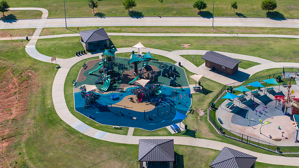 Aerial view of a circular playground with climbing structures, slides, shade canopies, connecting concrete paths, and small pavilion buildings surrounded by open green space.
