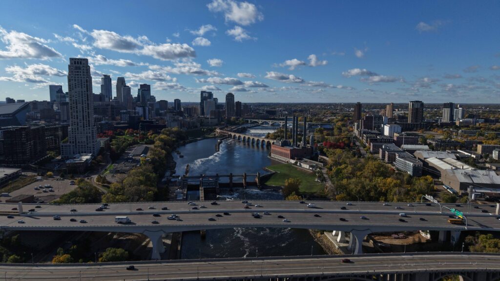 Aerial view of a river running through an urban area, with downtown buildings, bridges and highway traffic visible amid fall foliage.