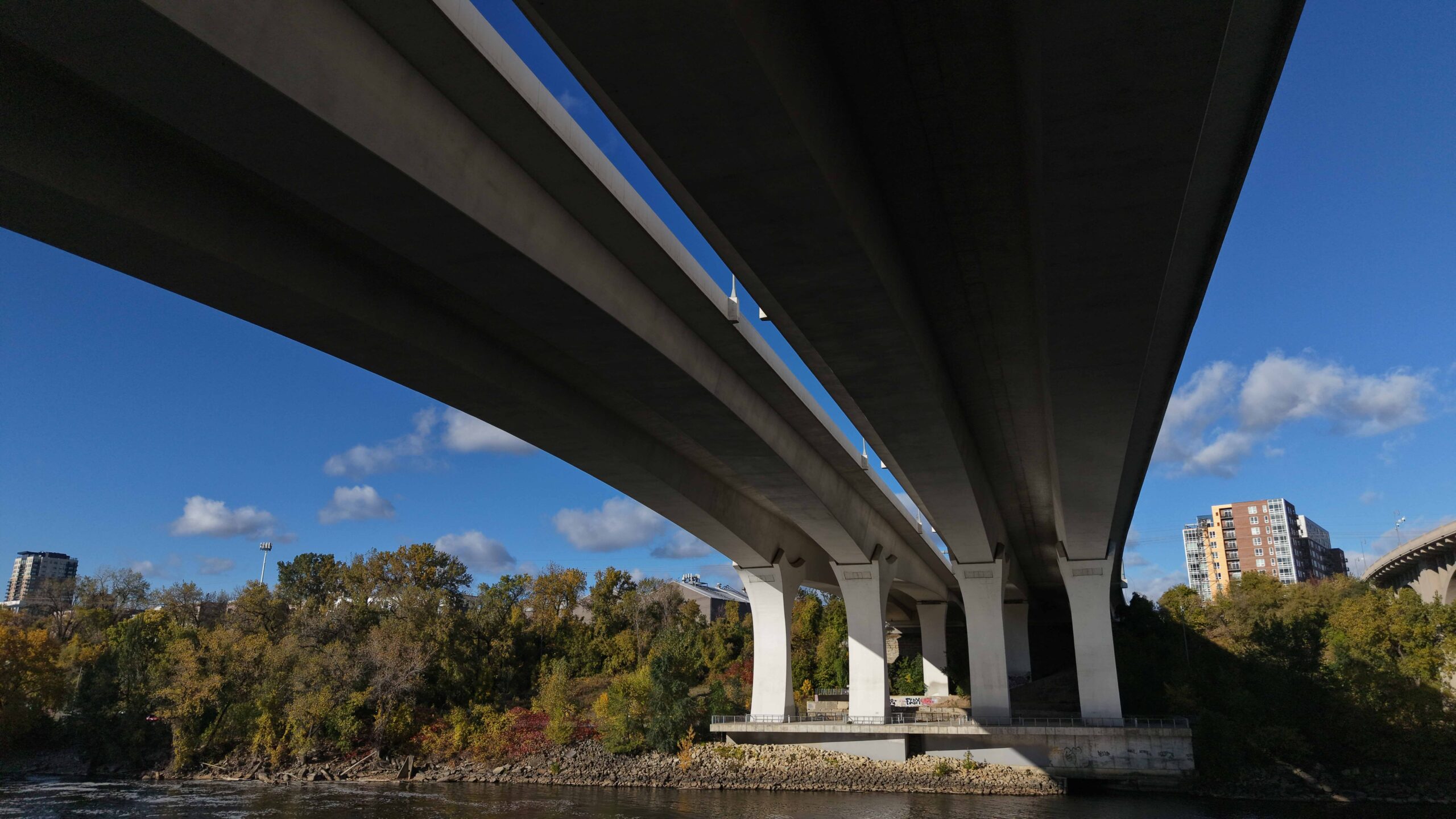 A view from beneath a multi‑span highway bridge crossing a river, showing concrete piers, the water below and tree‑lined riverbanks with buildings visible in the distance.