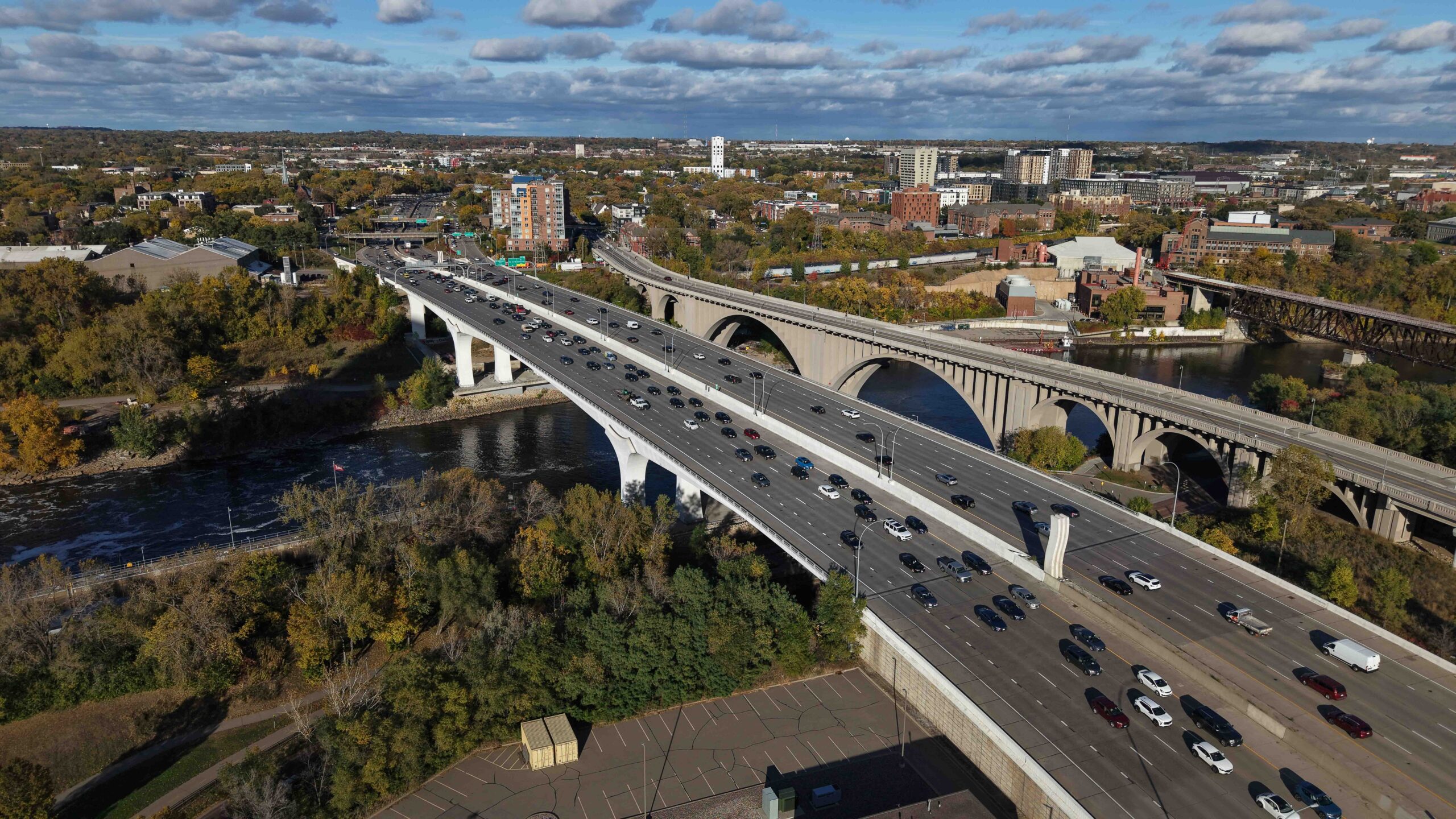 Aerial view of a wide interstate bridge carrying multiple lanes of traffic over a river, with adjacent historic arches, trees and nearby development.