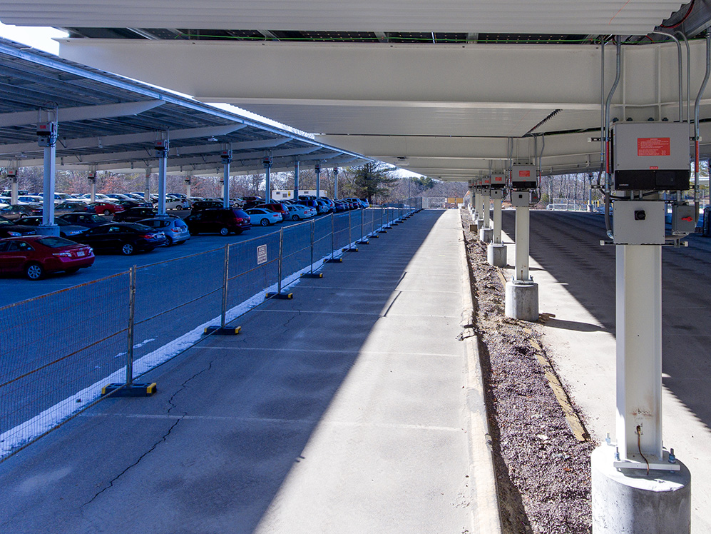View beneath solar carport structures at Stonehill College showing electrical equipment, conduit routing, and inverter installations along the parking area.