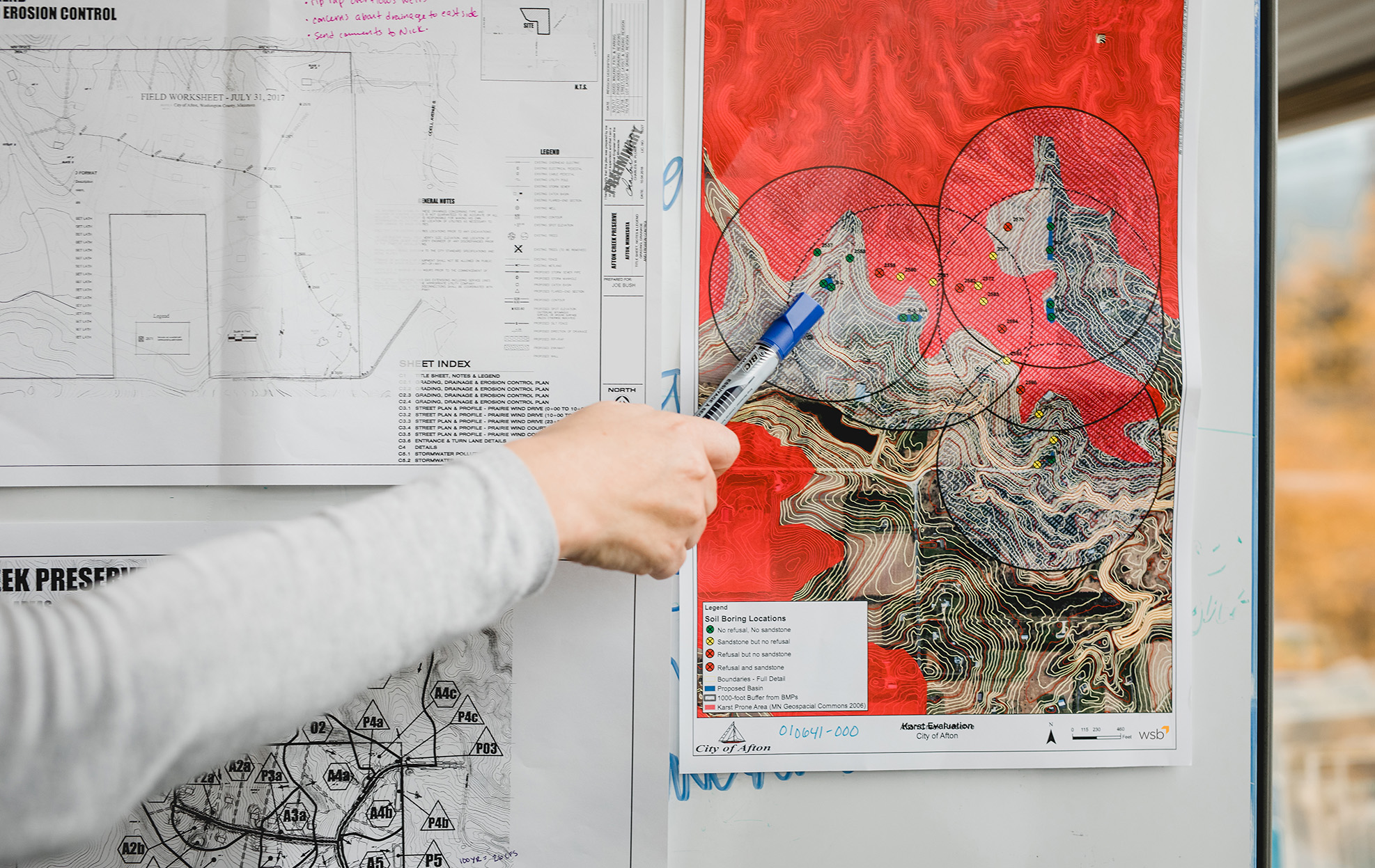Hand holding a pen points to a color‑coded erosion control map displayed alongside engineering plans on a wall.