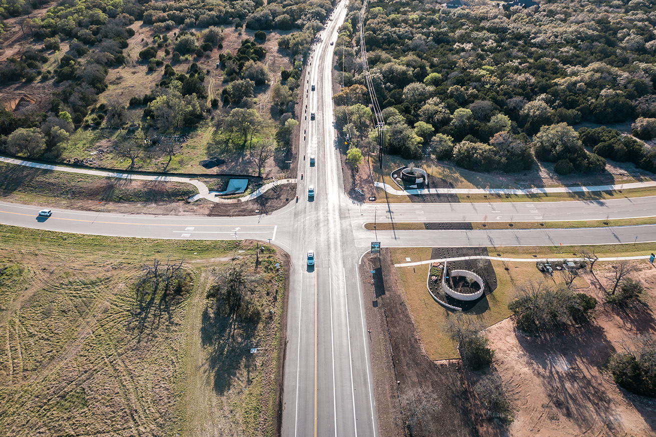 Aerial view of a multi-lane road crossing with cars, sidewalks, and landscaped paths surrounded by trees and green space.