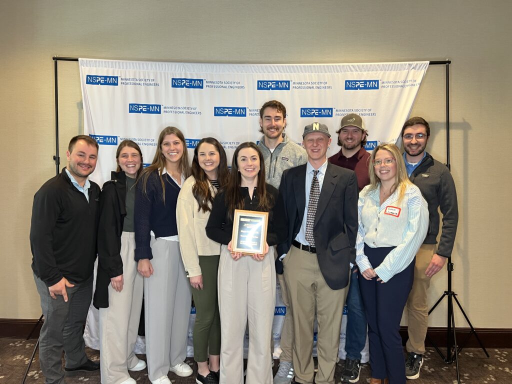 Group of professionals standing in front of an NSPE‑MN backdrop, with one individual holding an award plaque.
