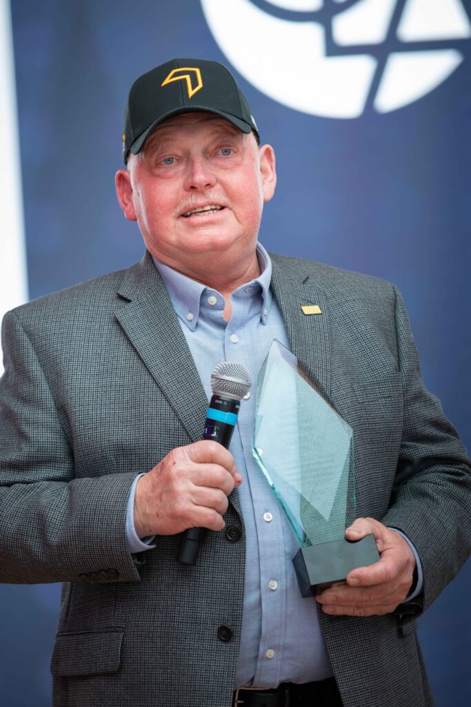 Close-up of Ron Bray holding a glass plaque and speaking into a microphone against a branded presentation backdrop.