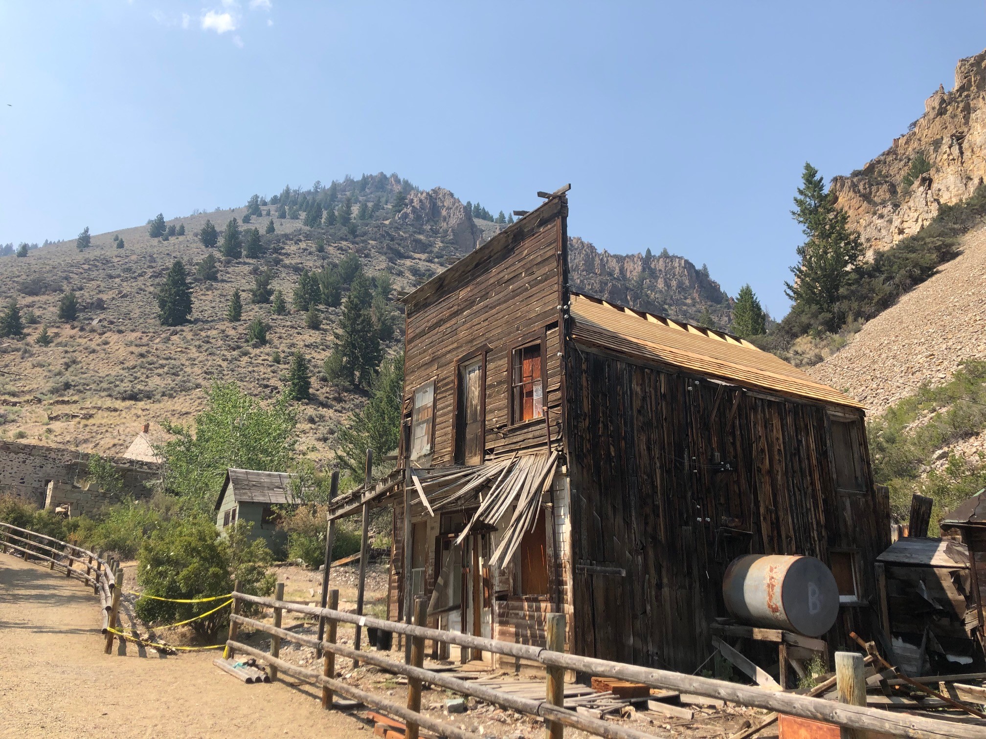 Weathered wooden mining building and remnants of historic structures at Bayhorse State Park with rocky hillsides and mountains in the background.