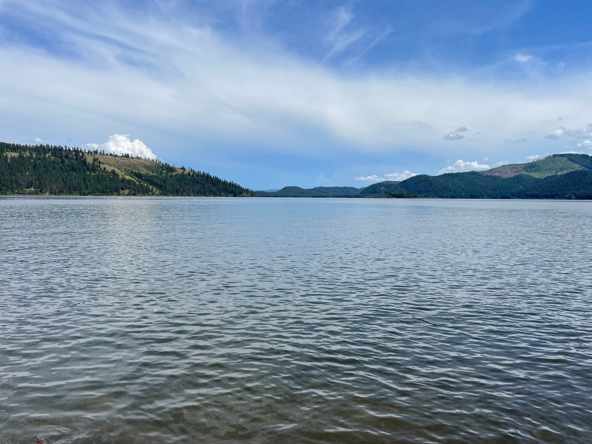 Wide view of a lake within the Coeur d’Alene River Basin surrounded by forested hills beneath a partly cloudy sky.