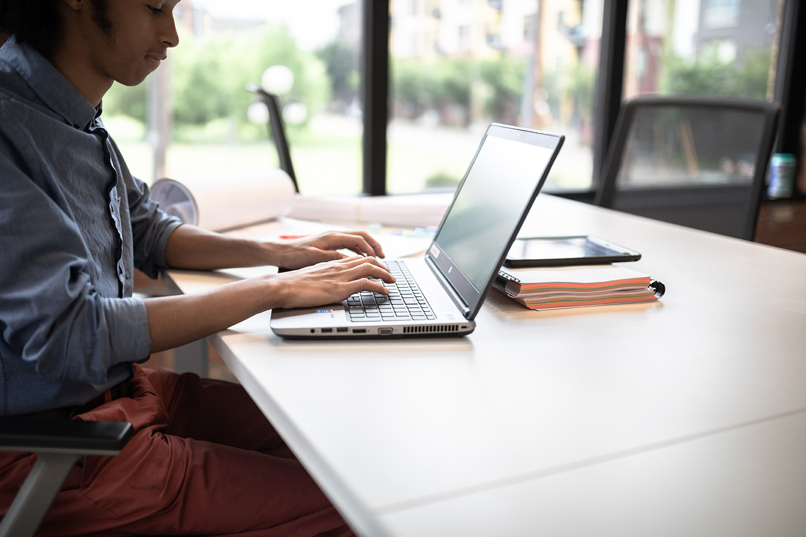 Person typing on a laptop at a desk in a bright office, with a tablet and a small stack of notebooks nearby; large windows in the background provide natural light.