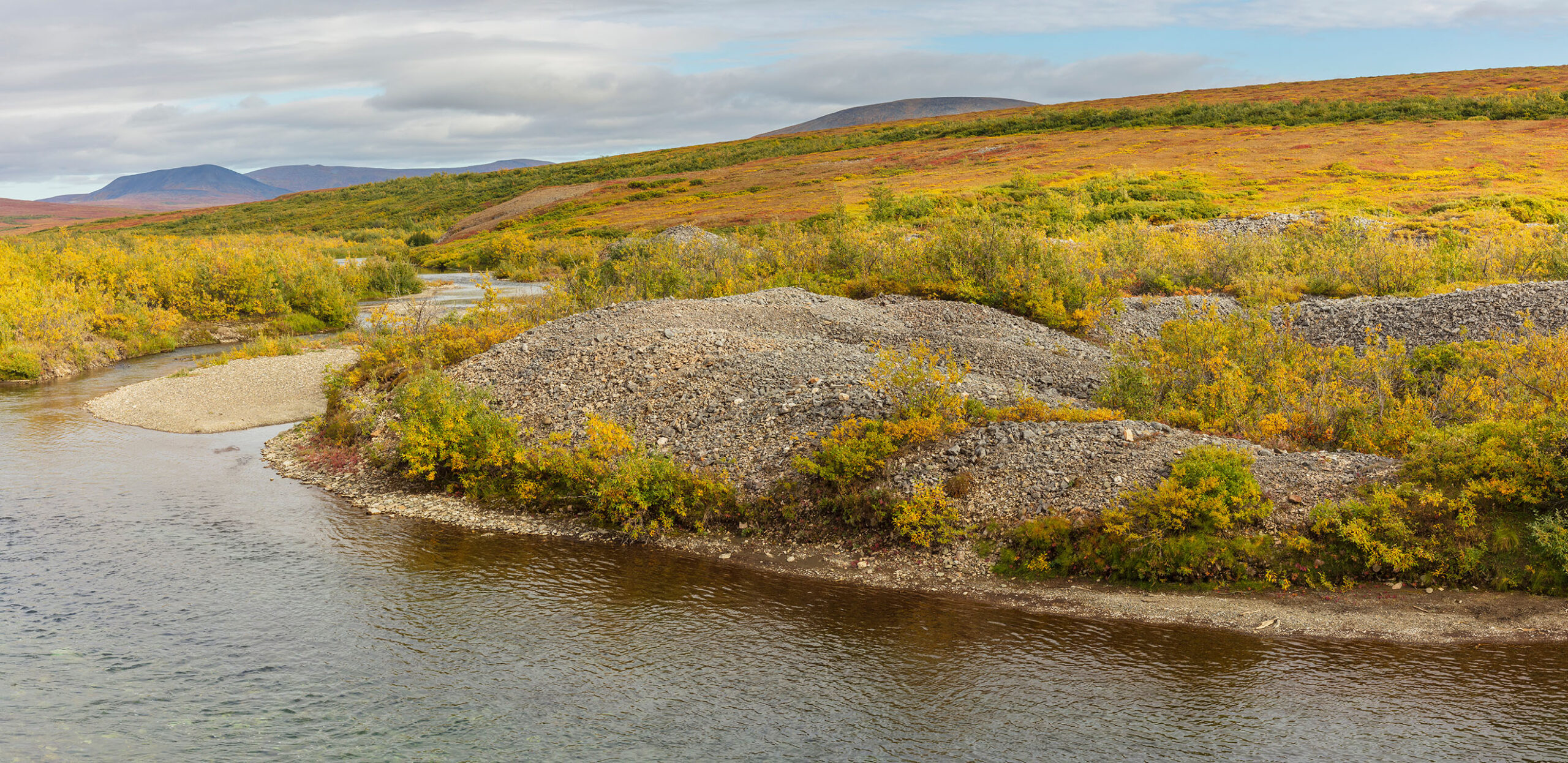 River winding through tundra landscape with gravel deposits, autumn vegetation, and rolling hills beneath a cloudy sky.