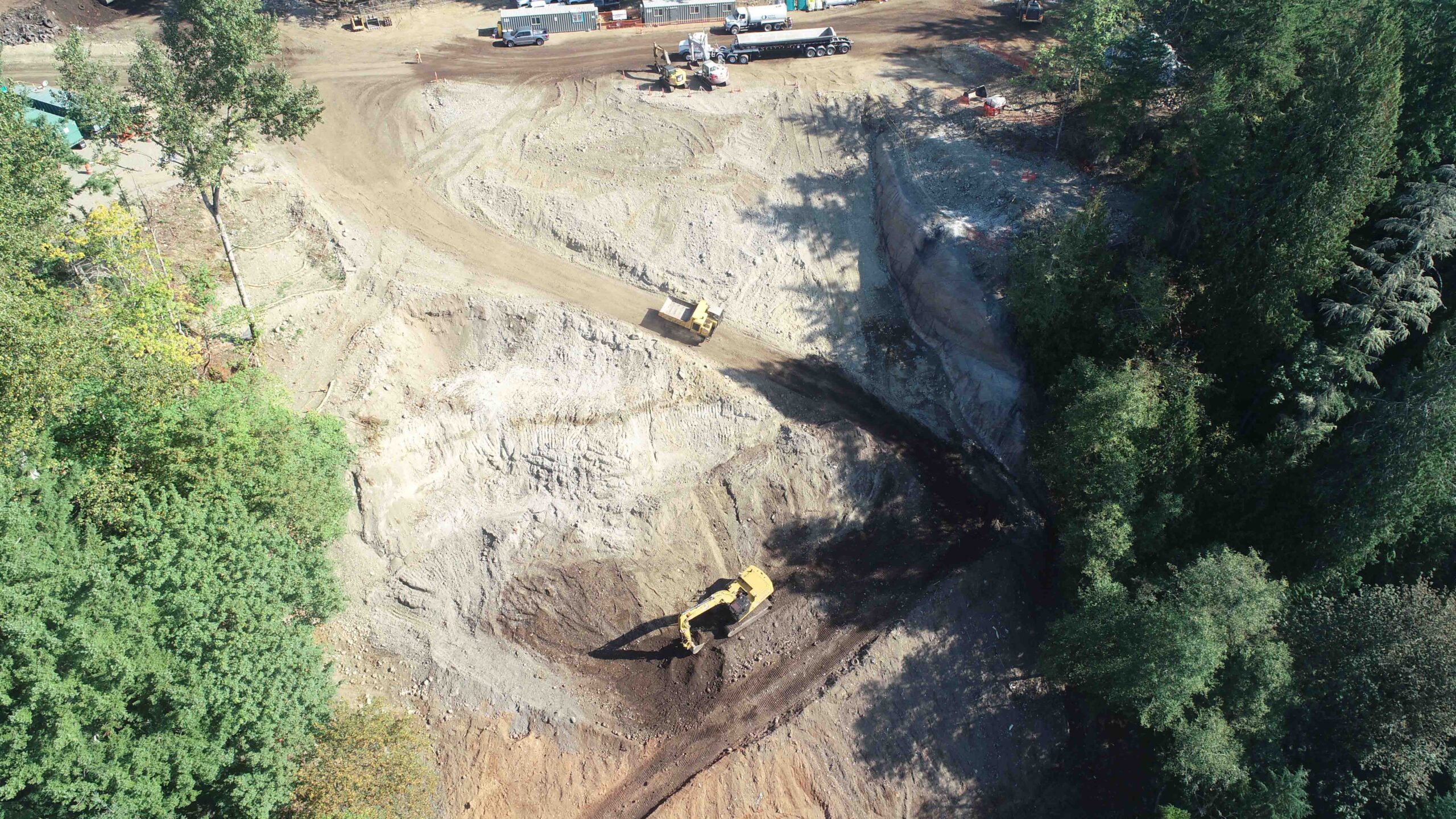 Aerial view of a large excavation area with heavy equipment operating on terraced slopes. An excavator and haul truck move material within the site, which is bordered by dense woodland and construction staging areas.