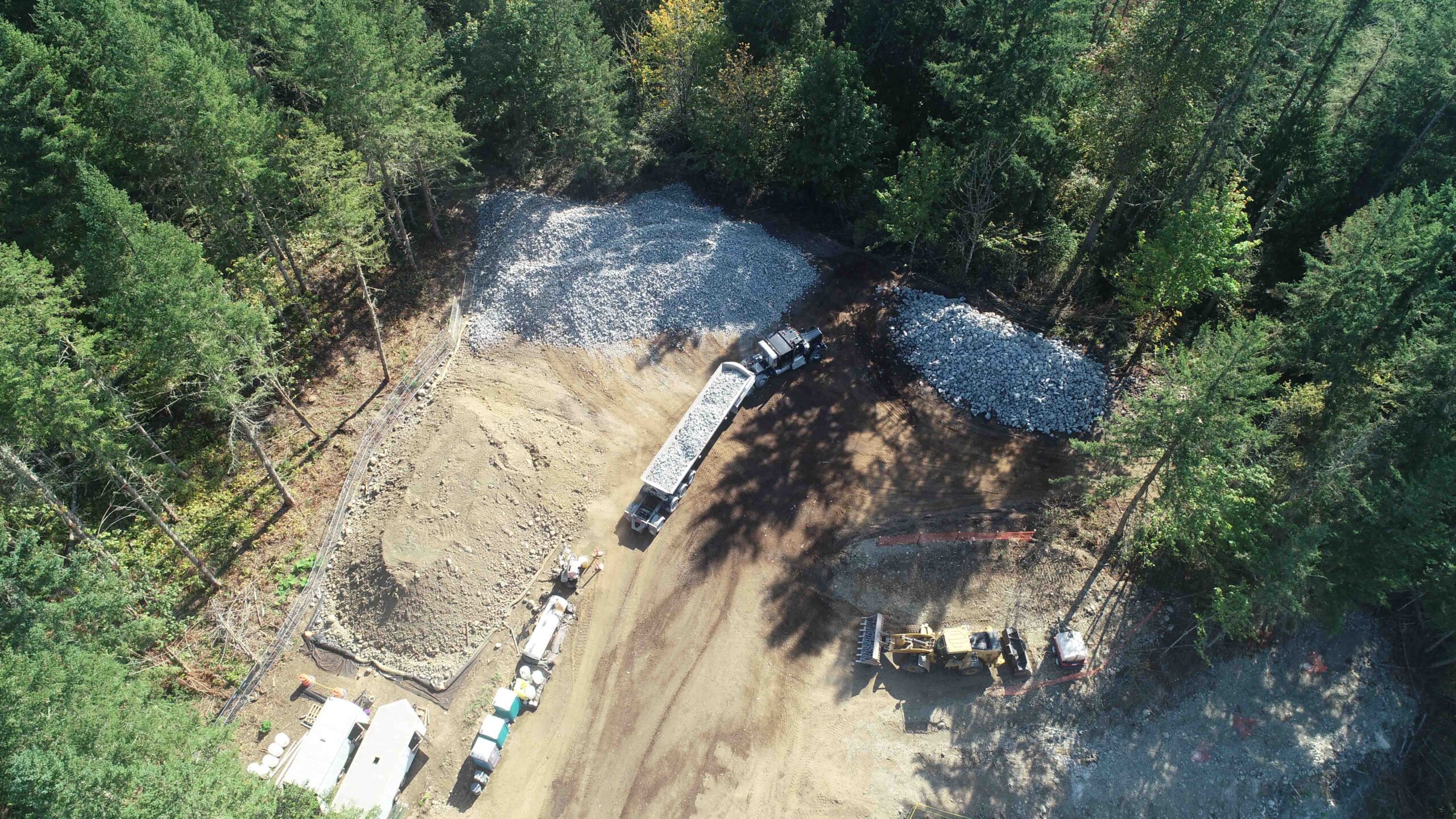 Aerial view of construction operations featuring a dump truck delivering rock material near large stockpiles. Heavy equipment and temporary site facilities are visible within a forested work zone.