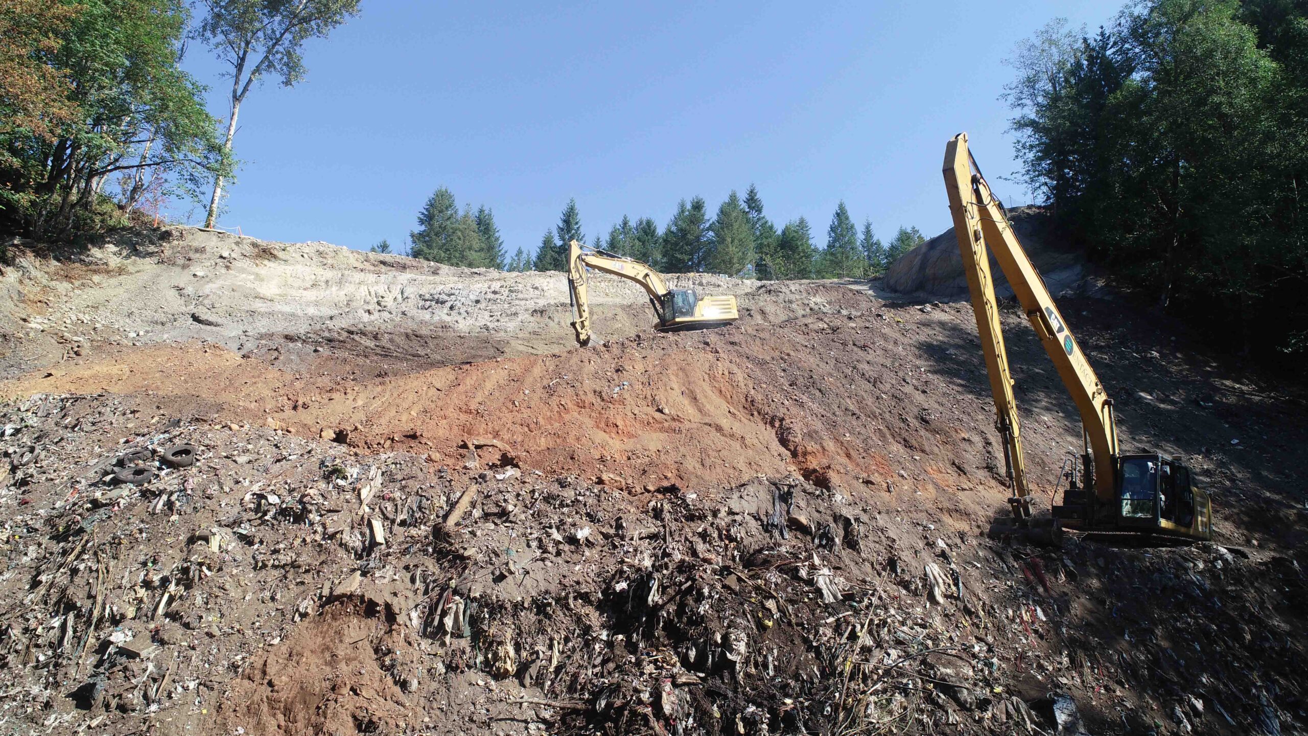 Excavators work on a steep excavation slope at a landfill remediation site, removing and relocating soil and waste materials. Forested hills surround the exposed earthwork area under a clear blue sky.