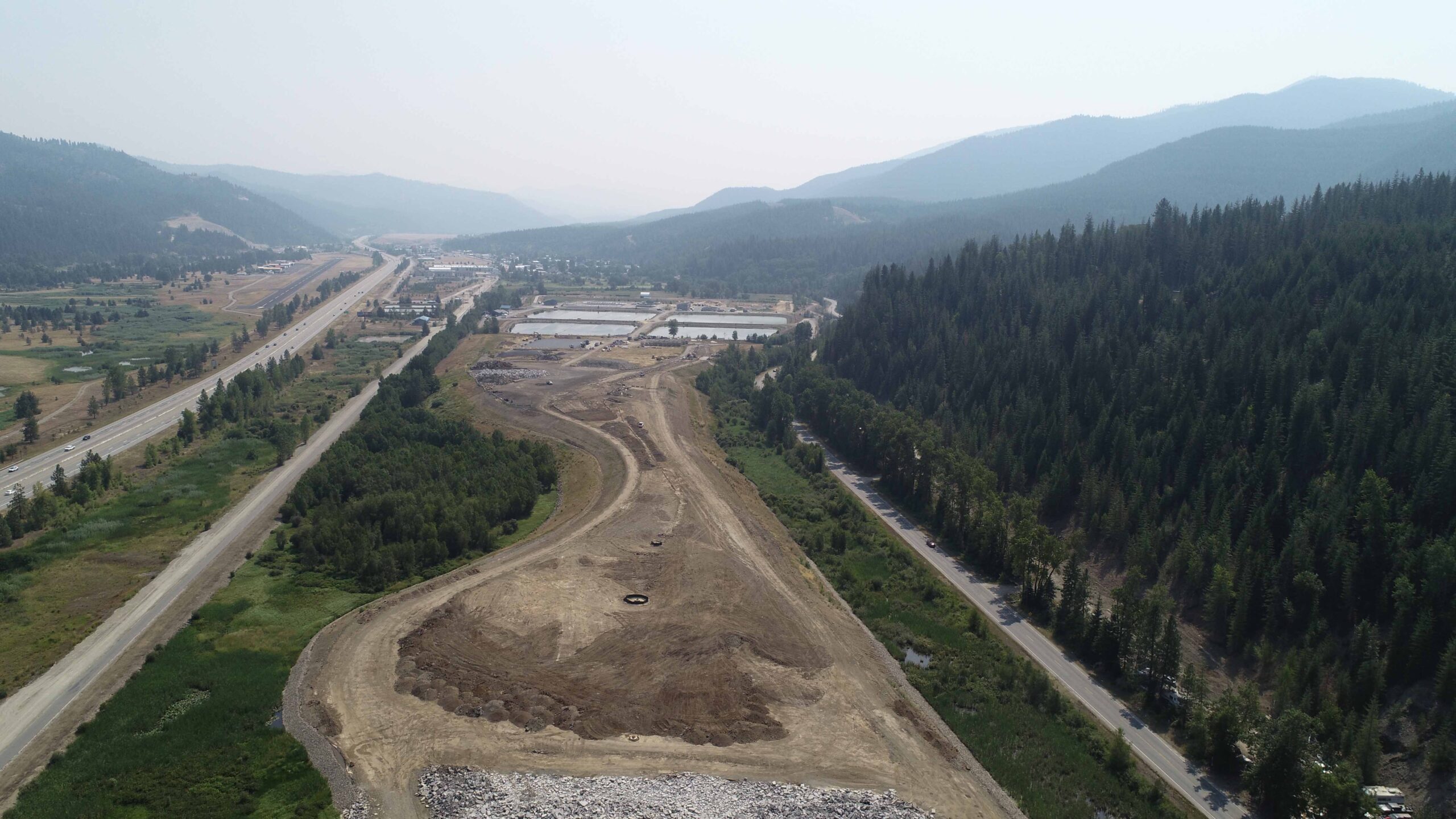 Wide aerial view of a large earthwork and environmental remediation site extending through a mountain valley. Access roads, treatment ponds, and graded land are visible alongside a highway and surrounding forested landscape.