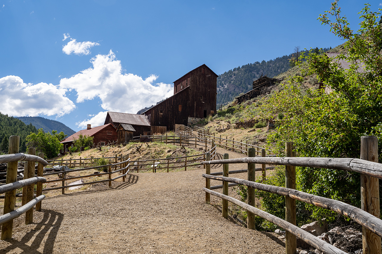 Historic mining buildings connected by a fenced walking path in a mountainous landscape under a bright blue sky.