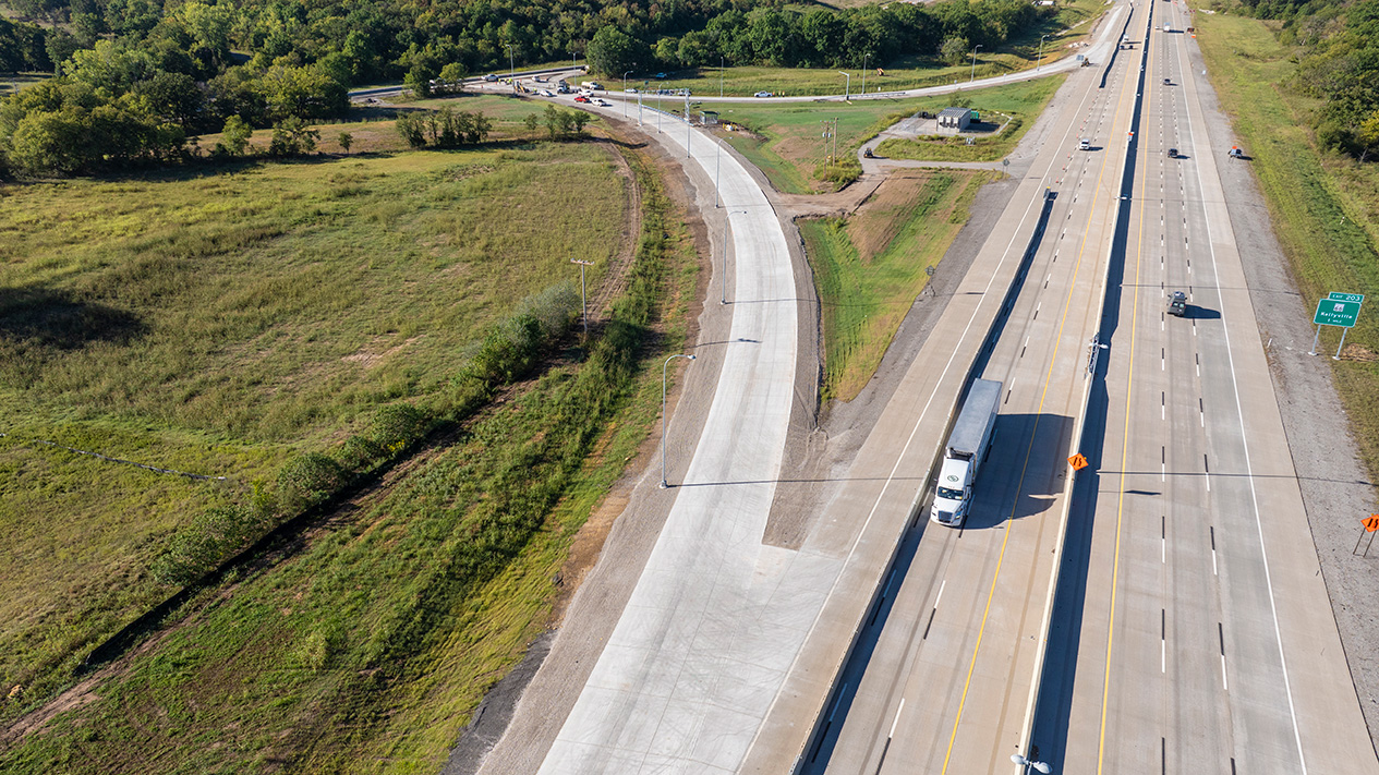 Aerial view of a highway interchange showing a newly constructed concrete ramp running parallel to a multi-lane interstate. Several vehicles, including a semi-truck, travel along the highway while grassy fields and drainage areas surround the roadway. Construction grading and roadway improvements are visible near the connecting ramp.