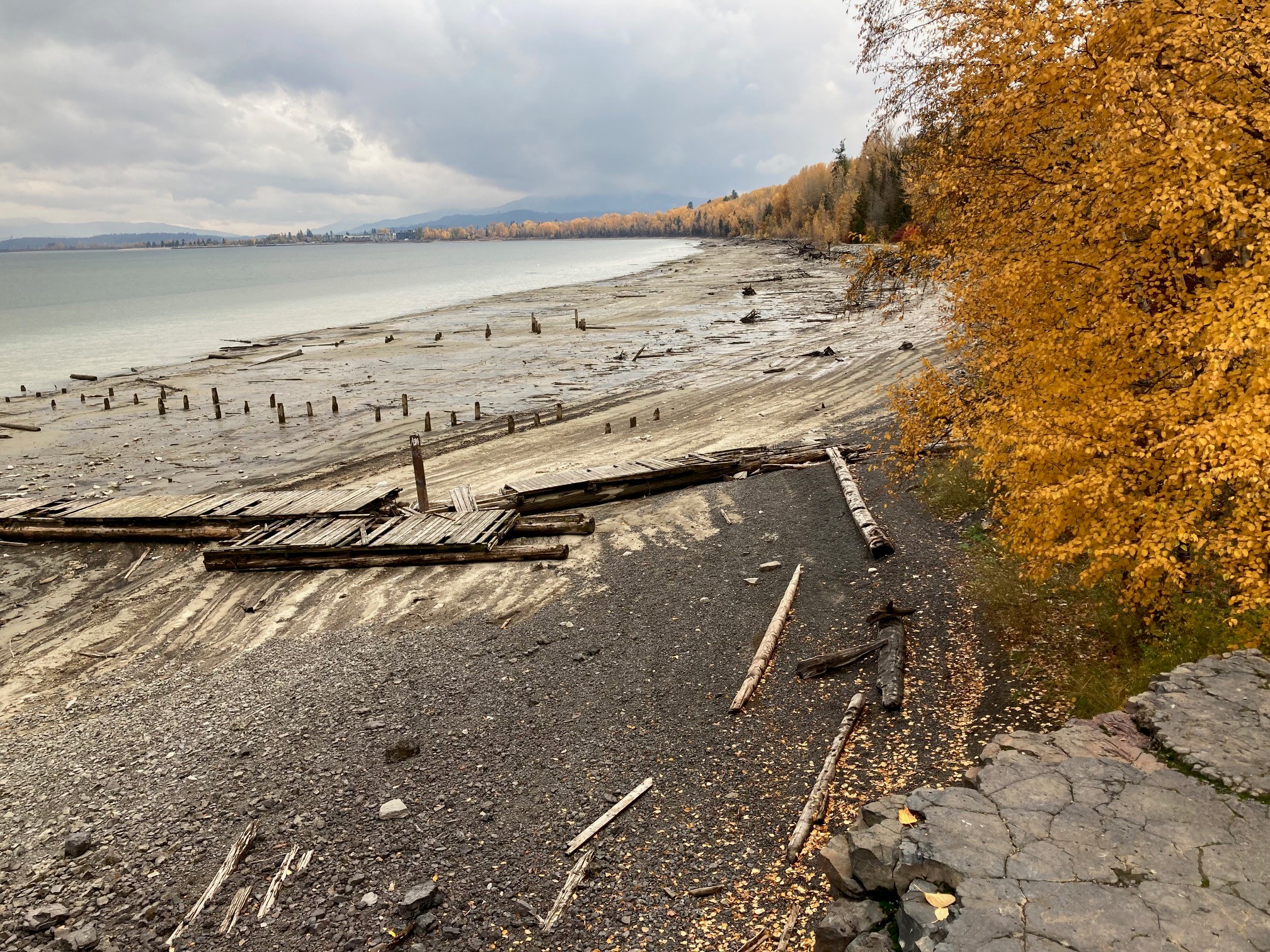 Exposed shoreline at Lake Pend Oreille with remnants of historic industrial structures, driftwood, and autumn foliage near the Ponderay Smelter Site.