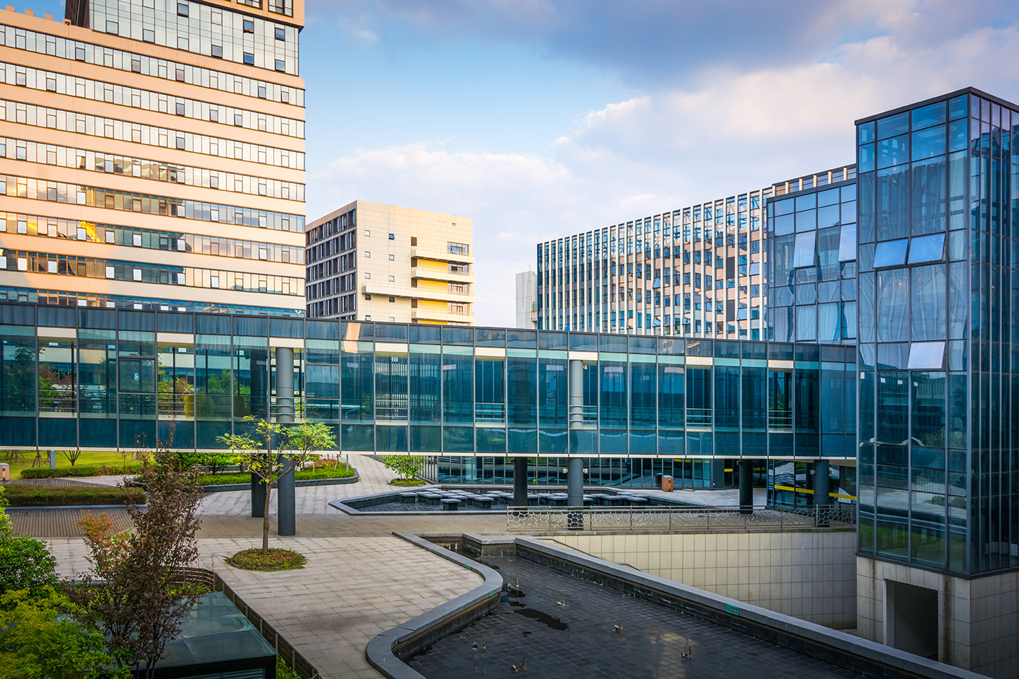 Modern glass and concrete buildings surrounding an open courtyard with landscaped walkways and reflecting pools.