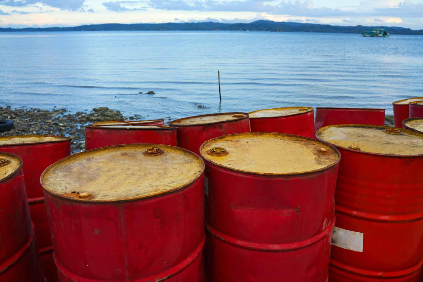 Red metal drums with weathered lids sit along a rocky shoreline, overlooking calm blue water and distant tree-covered hills.