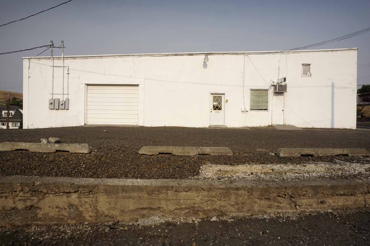 White industrial building beside a gravel lot and exposed concrete foundation.