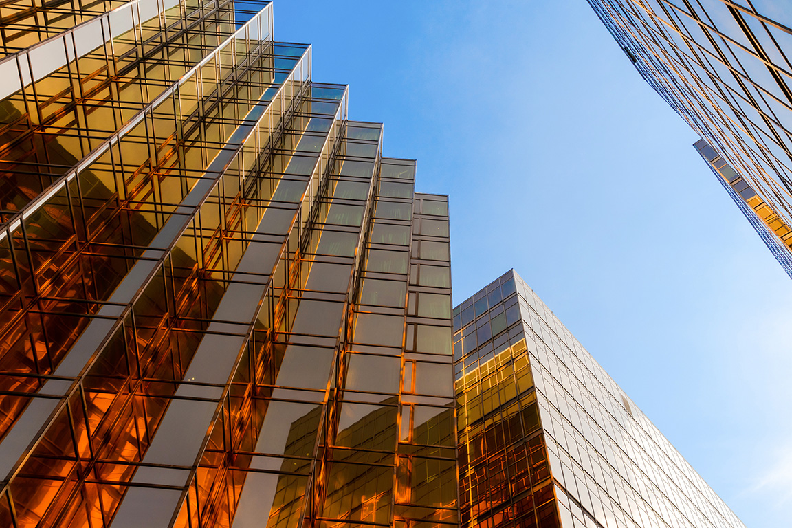 Low-angle view of modern glass office buildings reflecting warm sunlight against a clear blue sky.
