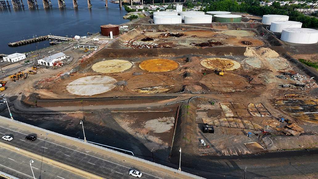 Aerial view of an industrial site under redevelopment beside a river, with cleared ground, circular concrete foundations, construction equipment and large storage tanks.