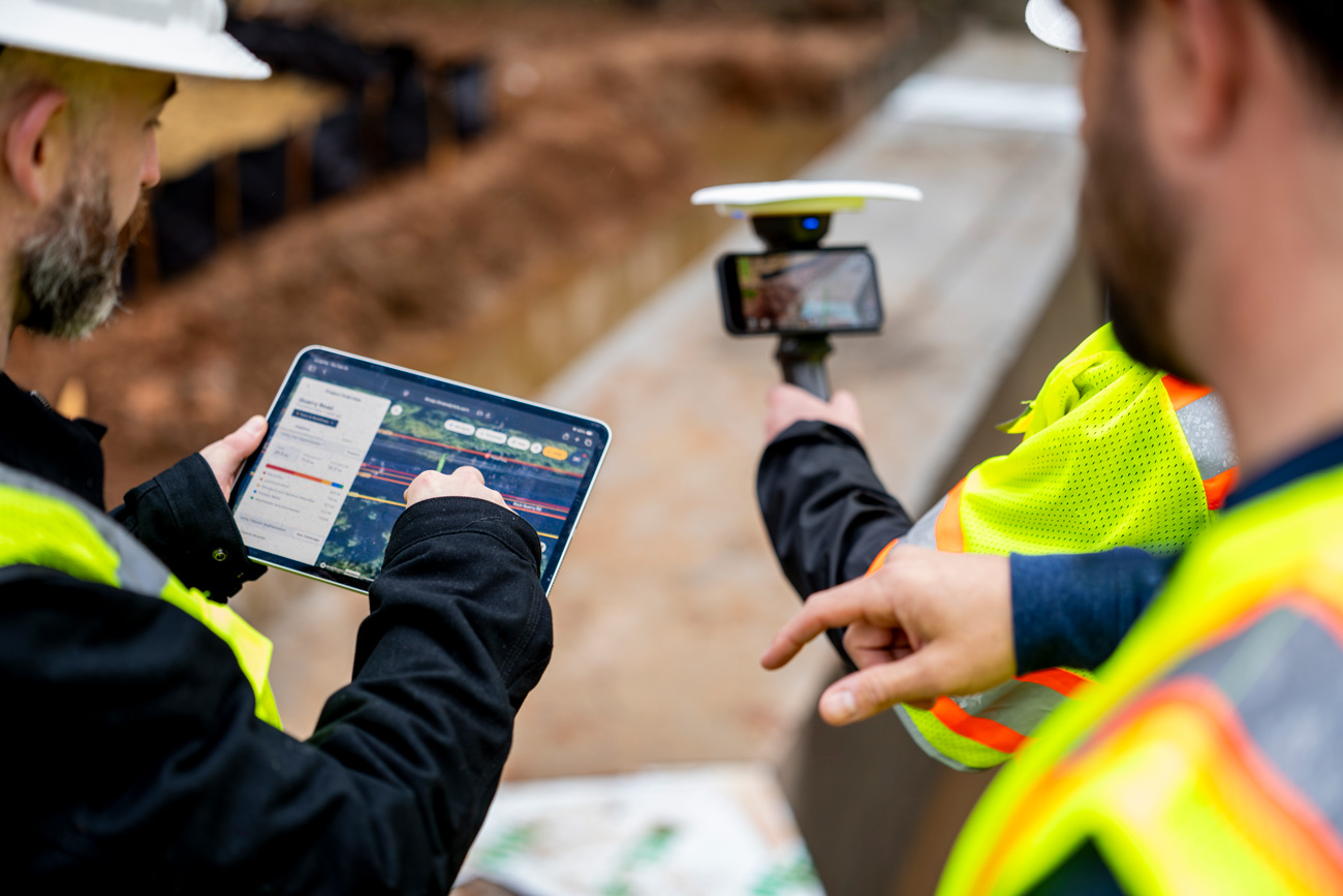 Construction surveyors in high-visibility vests discuss a digital site plan on a tablet while using a handheld scanner at a worksite, conveying teamwork.
