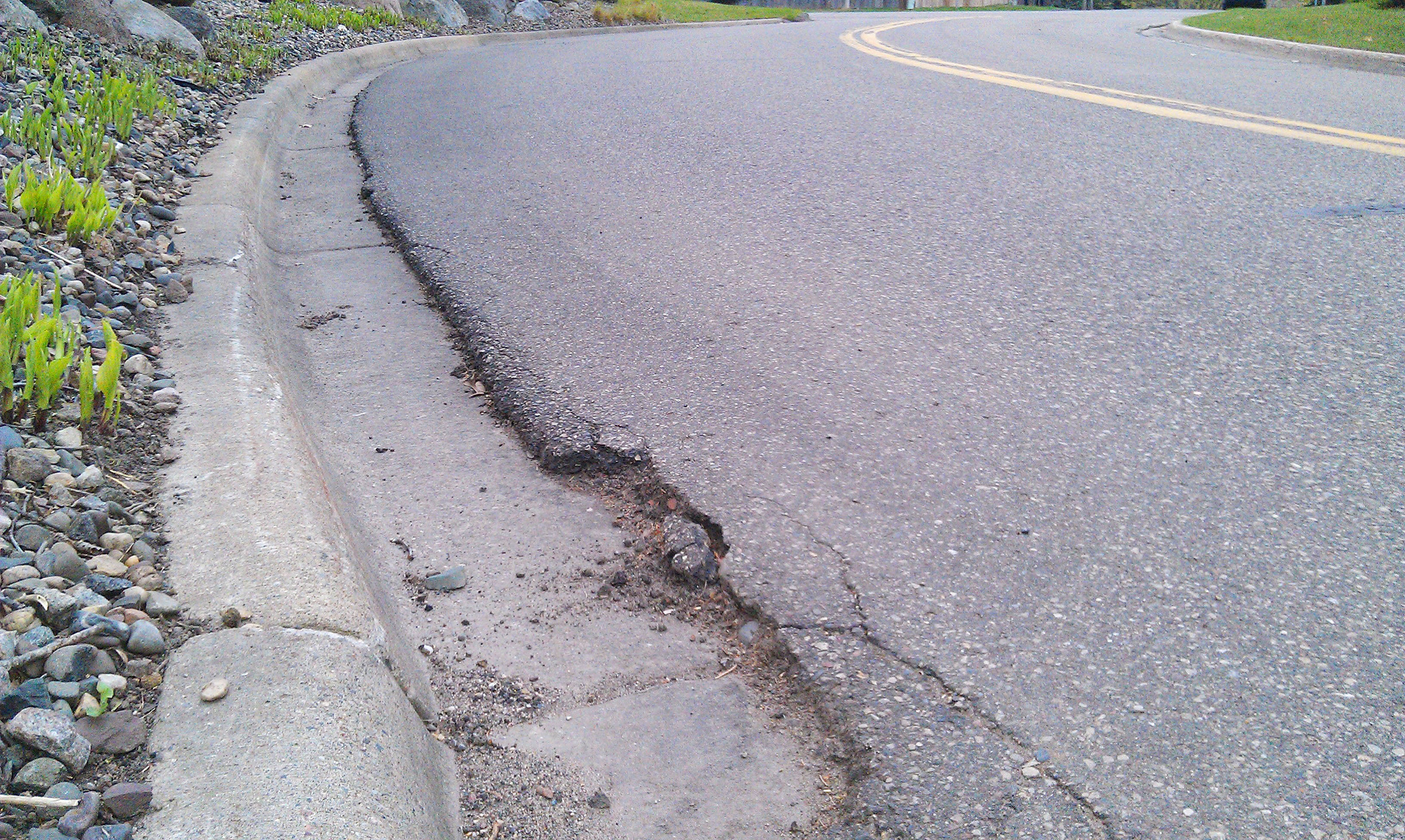 Cracked and deteriorating pavement along a concrete curb on a curved roadway.